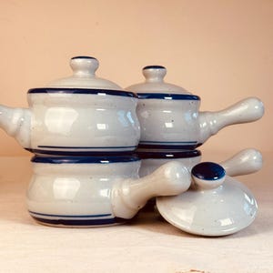 May include: A set of three off-white ceramic soup crocks with speckled textures and dark blue horizontal bands. Each crock features a single side handle and a matching lid with a small knob. One lid is placed separately in the foreground.