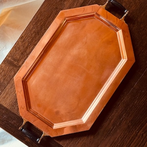 May include: An octagonal copper serving tray with a polished metallic finish and a raised border. The tray features two black handles attached to the ends with gold-toned metal hardware, providing a contrast to the warm copper surface.