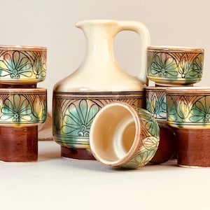 May include: A ceramic beverage set consisting of a central pitcher and several matching cups. Each piece features a cream-colored base with a brown bottom and a decorative band of green and brown floral-inspired patterns around the middle.
