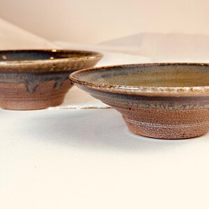 May include: Two ceramic bowls with a wide, shallow rim and a tapered base. The exterior features a textured, unglazed reddish-brown clay finish, while the interior and rim are coated in a smooth, glossy glaze with blended tones of blue, brown, and tan.