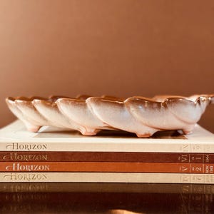 May include: A scalloped, light brown ceramic dish with a glossy finish, resting on a stack of four Horizon magazines. The dish features a repeating curved, petal-like edge design and small feet, showcasing a gradient of tan and cream tones.