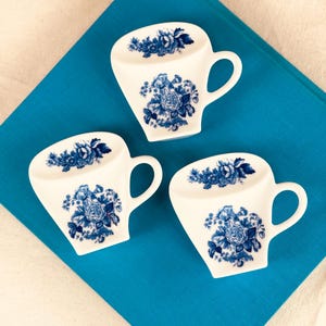 May include: Three white ceramic teacup-shaped coasters with blue floral designs. Each coaster has a handle and a flat surface decorated with a blue rose and other floral patterns. The coasters are arranged on a blue surface.