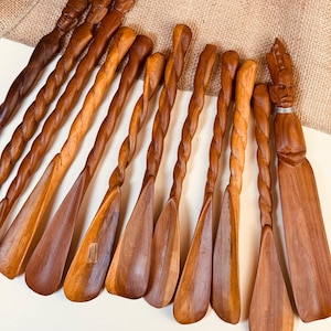 May include: A collection of ten wooden shoehorns arranged in a fan shape. Each shoehorn features a long, twisted handle and a smooth, wide scoop at the bottom. Several handles are topped with carved human head figures, and one has a small label.
