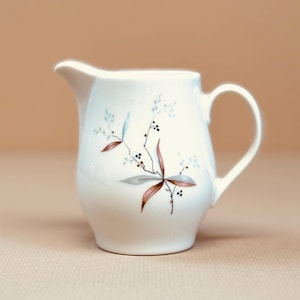 May include: A white ceramic creamer with a handle and spout. The creamer is decorated with a floral design in shades of brown, gray, and blue. The creamer is a classic design, perfect for serving cream or milk.