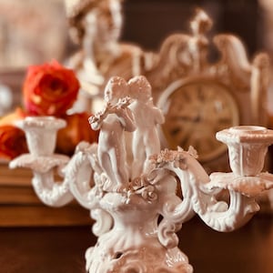 May include: White porcelain candle holder with three arms and a decorative design featuring two cherubs playing musical instruments. The base of the holder is adorned with floral details.