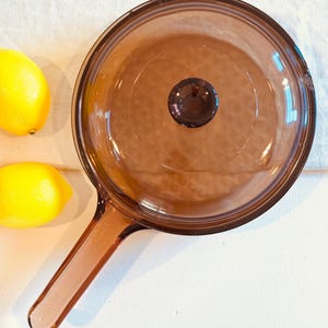 May include: A vintage amber-colored glass saucepan with a matching lid and handle. The lid has a dark brown knob. Two yellow lemons are visible in the background. The saucepan is on a white surface.