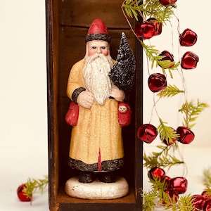 May include: A hand-painted Santa Claus figurine with a long white beard, wearing a cream-colored coat with a crackled finish, a red hat, and black boots. The figure holds a small black tree and stands inside a tall, narrow, dark wooden box.