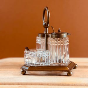 May include: An antique condiment set featuring a brass base and handle, with three clear glass containers. The containers have textured sides and are topped with brass lids. A small spoon rests in one container.
