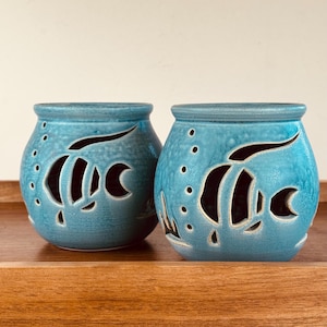 May include: Two rounded ceramic tealight holders in a vibrant turquoise blue glaze. Each vessel features a cut-out design of a tropical fish surrounded by small circular bubbles, allowing light to pass through the open spaces in the ceramic walls.