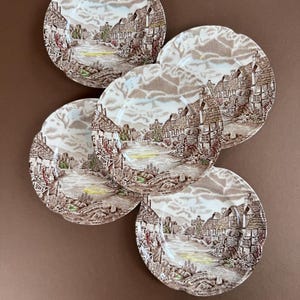 May include: A set of five round ceramic plates featuring a brown and white transferware pattern of a village scene with cottages, a river, and trees. Each plate has a scalloped edge and a detailed landscape illustration printed on the surface.