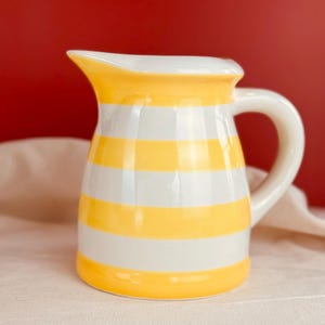 May include: A ceramic pitcher with a white handle and spout. The pitcher is decorated with horizontal yellow and white stripes. The background is a solid red color.