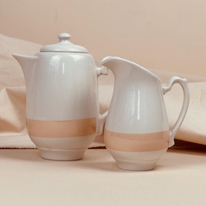May include: A white ceramic teapot with a lid and a matching creamer. Both pieces have a horizontal peach-colored band around the middle. The teapot has a spout and handle, while the creamer has a handle and a pouring lip.