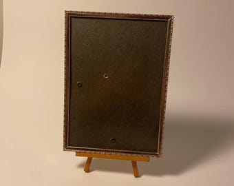 May include: A silver picture frame with a decorative border sits on a small wooden easel. The frame is empty and ready for a photo.