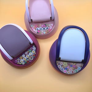 Peut inclure: Trois supports de téléphone personnalisés colorés. Les supports sont dans des tons de rose, de violet et de prune, chacun avec une poignée blanche. Les bases comportent des noms épelés avec des accents floraux et des visages souriants. Le support central porte le nom "LUBA".