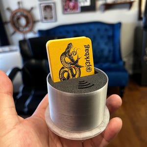 May include: A yellow business card with a black snake illustration and the text "@j3rkbag" is displayed in a silver card holder. The card holder is held in a person's hand. The background includes framed artwork and a blue chair.