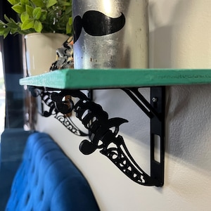 Może przedstawiać: Turkusowa półka z czarnymi metalowymi wspornikami w kształcie sztyletów. Półka jest zamontowana na białej ścianie i ma srebrny kubek z czarną naklejką z wąsem.