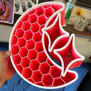 May include: A crescent moon and star-shaped wall shelf featuring a bright pink interior with a honeycomb grid pattern and a solid white outer frame. The structure includes multiple hexagonal compartments and two larger star-shaped storage sections.