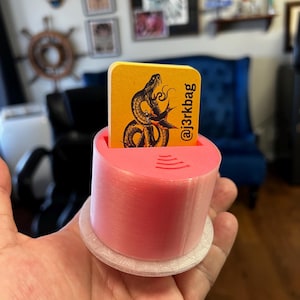 May include: A pink 3D-printed card holder with a white base, holding a yellow card. The card features a black snake illustration and the text "@j3rkbag". The holder has a contactless payment symbol.