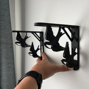 May include: Two black metal shelf brackets with a swallow design. The brackets are mounted on a white wall.