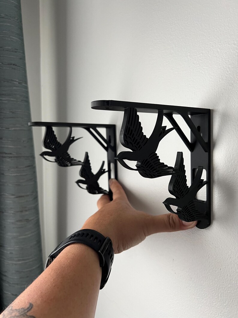 May include: Two black metal shelf brackets with a swallow design. The brackets are mounted on a white wall.