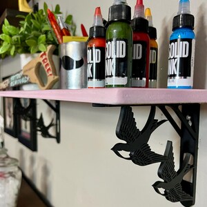 May include: A pink shelf with black swallow-shaped brackets. The shelf is holding six bottles of tattoo ink. The bottles are labeled "SOLID INK" and are in the colors green, red, orange, and blue.