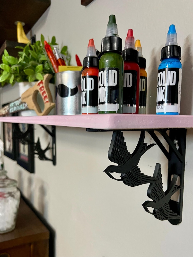 May include: A pink shelf with black swallow-shaped brackets. The shelf is holding six bottles of tattoo ink. The bottles are labeled "SOLID INK" and are in the colors green, red, orange, and blue.