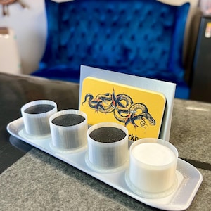 May include: A white tray with four clear plastic containers, three filled with black ink and one filled with white ink. The tray is on a black surface. A business card with a yellow background and a black snake tattoo design is behind the tray. The business card says "remarkable".