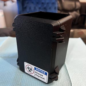 Peut inclure: Un conteneur noir pour l'élimination des objets tranchants avec une surface texturée. Le conteneur a un couvercle à charnière et une étiquette avec un symbole de risque biologique et le texte "NOTICE Sharps disposal."