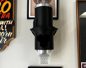 May include: A black, cylindrical tattoo machine holder with a silver top and a hexagonal design. The holder is mounted on a wall, with framed artwork visible in the background.