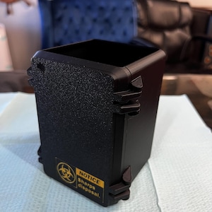 Op de afbeelding: Een zwarte, rechthoekige container voor het weggooien van scherpe voorwerpen met een getextureerd oppervlak. De container heeft een geel en zwart biohazard-symbool en de tekst "NOTICE Sharps disposal."