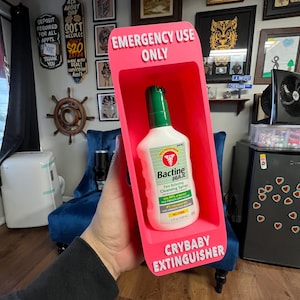 May include: A pink plastic holder labeled "EMERGENCY USE ONLY" and "CRYBABY EXTINGUISHER" contains a bottle of Bactine MAX pain-relieving cleansing spray. The holder is being held up by a person. The background includes framed art and a blue chair.