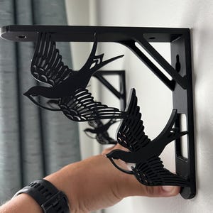 May include: Two black metal shelf brackets with a swallow design. The brackets are mounted on a white wall.
