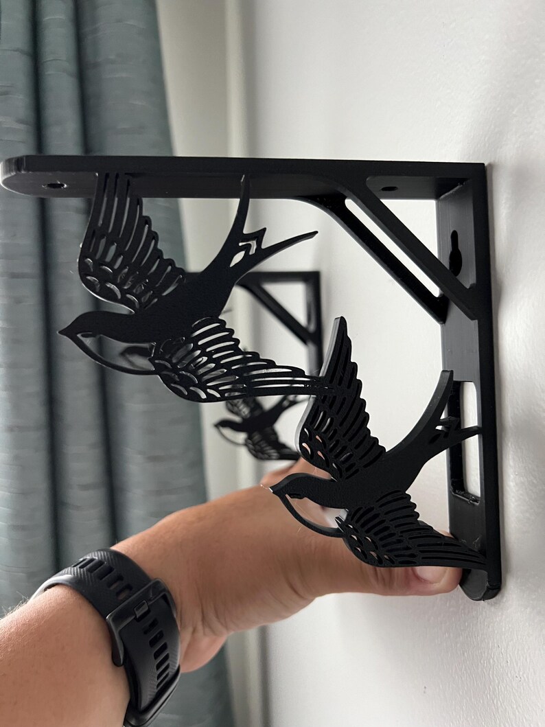 May include: Two black metal shelf brackets with a swallow design. The brackets are mounted on a white wall.