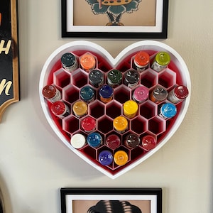 May include: A heart-shaped, white and red organizer filled with colorful ink bottles. The organizer has a honeycomb design and is mounted on a wall. The image also shows framed artwork.