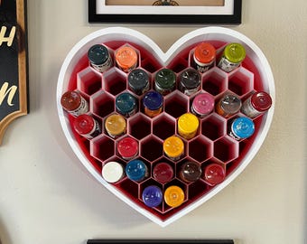 May include: A heart-shaped organiser in white and red, filled with various coloured ink bottles. The organiser has a honeycomb design and is mounted on a wall. Framed artwork is also visible.