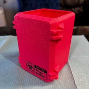 Op de afbeelding: Felroze bak voor het weggooien van scherpe voorwerpen. De bak heeft een getextureerd oppervlak en een biohazard-symbool met de woorden "NOTICE Sharps disposal". De bak is rechthoekig met een vierkante opening.