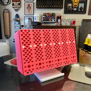May include: A pink desktop organiser featuring five vertical compartments with a repeating heart-shaped cutout pattern on the front panels. The organiser is supported by a white rectangular base and includes small white heart accents on each section.