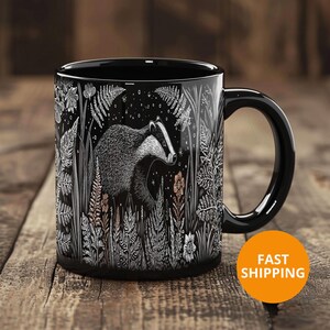 May include: Black ceramic mug featuring a detailed illustration of a badger in a forest setting. The design includes ferns, flowers, and stars, all in white against a black background. The mug has a curved handle and is set on a wooden surface.