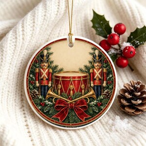 May include: A round ceramic ornament featuring a festive illustration of two nutcracker soldiers standing on either side of a red drum, with crossed gold trumpets, a large red bow, and evergreen branches, all suspended by a gold string.