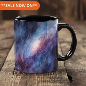 May include: Black ceramic mug with a watercolour galaxy design in blue, purple, and white. The mug has a black handle and rim. The text "SALE NOW ON" is in an orange banner.