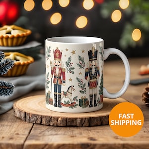 May include: A white ceramic mug featuring a festive pattern of two nutcracker figures, a rocking horse, wrapped gifts, pine branches, and gold stars. One nutcracker wears a red uniform, while the other wears a dark blue uniform with a drum.