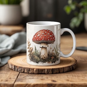 May include: White ceramic mug featuring a detailed illustration of a red and white spotted mushroom. The mushroom is set against a backdrop of ferns and foliage, creating a whimsical, nature-inspired design. The mug is placed on a wooden coaster.