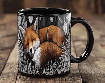 May include: Black ceramic mug featuring a sleeping fox illustration. The fox is depicted with orange and white fur, nestled among detailed black and white foliage. The mug has a black handle and a glossy finish, perfect for coffee or tea.