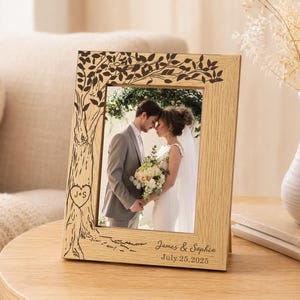 May include: A rectangular light wood picture frame featuring a laser-engraved tree design on the border, a heart with the initials J+S, and the text James & Sophia July 25, 2025. The frame holds a photograph of a couple in wedding attire.