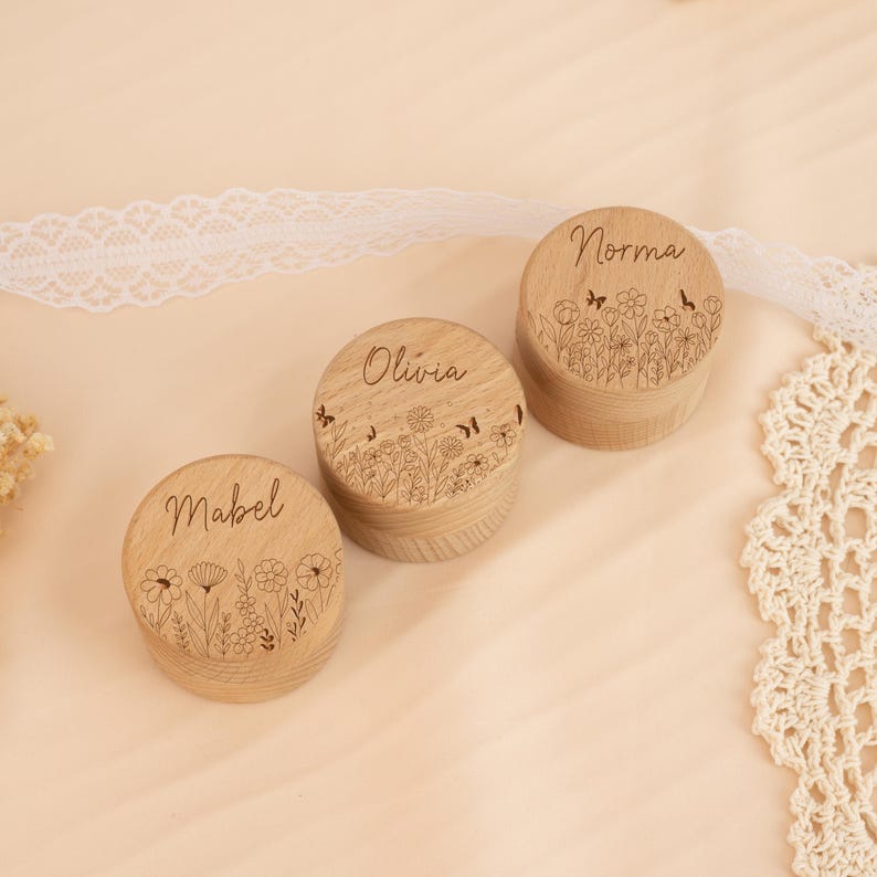 May include: Three round wooden boxes with floral designs and names engraved on the top. The names are Mabel, Olivia, and Norma.