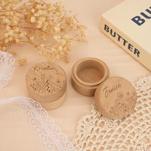 May include: Two wooden ring boxes with floral designs and the names "Kristin" and "Janice" engraved on them. The boxes are on a white lace doily and a beige surface.