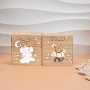 May include: Two wooden money boxes with slots on top. One features a pink elephant on a cloud with the name "Sophia" and stars. The other has a teddy bear with the name "Daniel" and stars. Both are light brown.