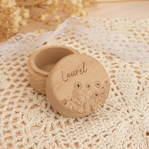 May include: A small wooden box with a lid, engraved with the name "Laurel" and a floral design. The box is light brown and has a natural wood finish.