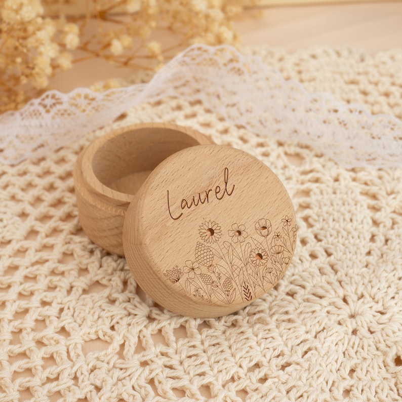 May include: A small wooden box with a lid, engraved with the name "Laurel" and a floral design. The box is light brown and has a natural wood finish.