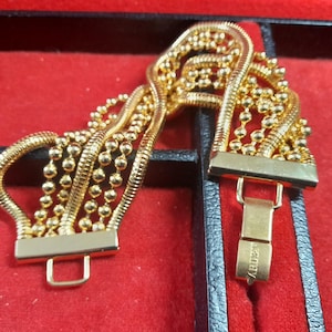 Könnte beinhalten: Ein goldfarbenes Armband mit mehreren Strängen aus kleinen, runden Perlen und dickeren, gebogenen Bändern. Das Armband wird auf einer roten Oberfläche in einer schwarzen Box präsentiert, was sein aufwendiges Design und seinen metallischen Glanz hervorhebt.
